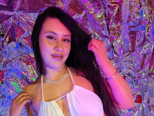CeleneEvans with long black hair, white halter top, showing her tits, holding hair, smiling, purple and blue light, silver necklace, and bracelet, against shiny metallic backdrop.