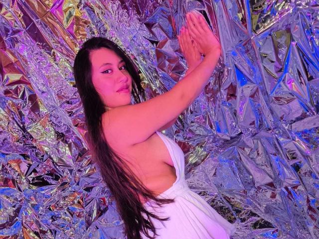 CeleneEvans with long black hair, wearing a white dress with a deep side slit, raising her hands against a shiny, metallic background. Her tits are partially visible.