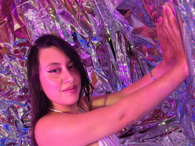 CeleneEvans with pink eyeliner, long dark hair, and a lip piercing, smiling while pressing against shiny, crumpled foil with purple and pink lighting. Boobs visible, arms raised.