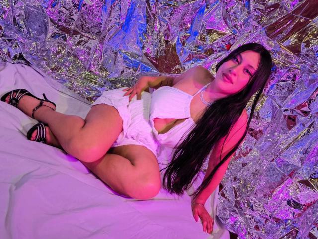 CeleneEvans with long black hair, wearing a white halter top and shorts, showing her ass and small tits, sitting on a white sheet, in black strappy heels.