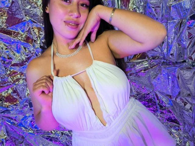 CeleneEvans with large boobs, wearing a white halter dress, touching her chin, smiling, long brown hair, silver necklace, purple and blue lighting, standing against crinkled silver foil.