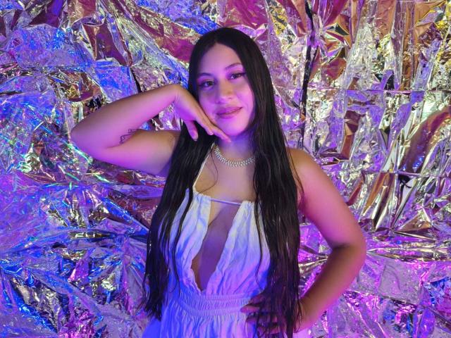 CeleneEvans with long black hair, smiling, wearing a white dress with a deep V-neck, silver necklace, hand on cheek, standing against shiny metallic background with purple and blue lights.