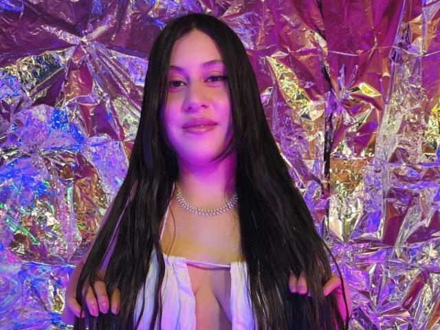 CeleneEvans with long black hair, purple light, white low-cut top, visible cleavage, necklace, holding hair, smiling, silver foil background, colorful reflections.