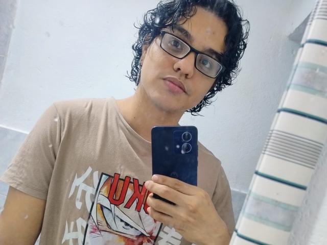 Diane69, con capelli neri ricci, occhiali e una maglietta beige, tiene in mano un telefono blu, sta scattando un selfie allo specchio, mostrando un'espressione neutra. Seno: nessuno, sedere: nessuno, tette: nessuno, vagina: nessuno..