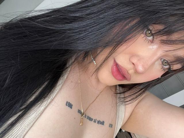 DanielaRuizz with long black hair, green eyes, and pink lips, wearing a beige tank top, showing her small tits and tattooed chest, wearing a gold necklace.