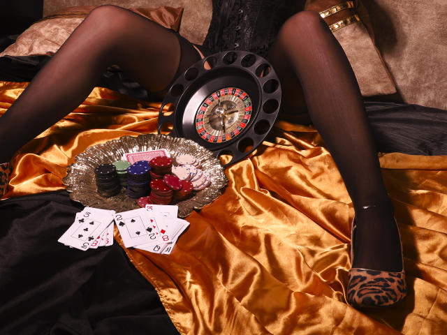 NoemyAshe's legs spread, wearing black stockings and leopard heels, with a poker game on a golden satin bed, featuring chips, cards, and a clock. Pussy not visible.