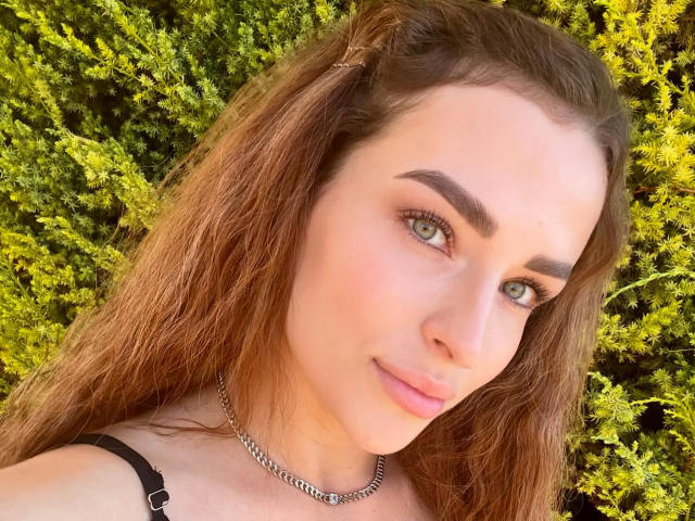MandiBrooks has light green eyes, brown wavy hair, and a subtle smile, wearing a black strap and a silver chain, with a clear, smooth complexion.