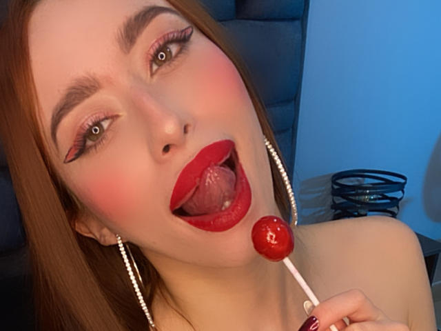 SarahLuxe with red lipstick, tongue out, holding a red candy on a white stick, has green eyes, pink eyeshadow, and long brown hair. She's wearing pearl earrings.