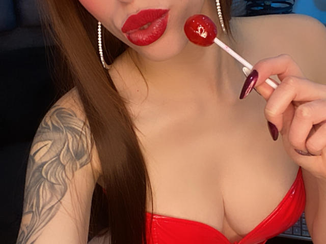 SarahLuxe with red lipstick, holding a red lollipop near her glossy pink lips, wearing a red bra that highlights her big tits, and showing a tattoo on her right arm.