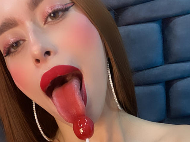 SarahLuxe with red lipstick, tongue out, and a red candy in her mouth, wearing silver earrings, has flushed cheeks and brown hair, looking seductive.