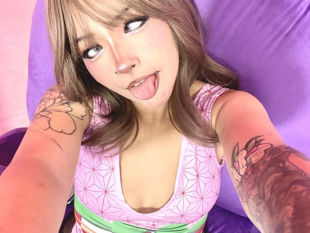 CarlaMiceli with brown hair, blue eyes, nose ring, and tongue out, wearing a pink geometric top, showing tattoos on arms, and a green waistband, taking a selfie.