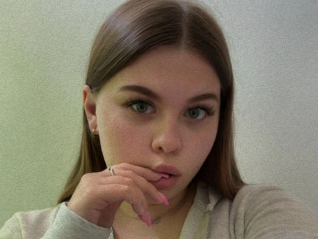 AbbyMaus-hot with brown hair, green eyes, pink nails, and a hand near her mouth, wearing a beige top, looking at the camera. Her nose is slightly upturned.