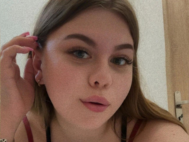 AbbyMaus-hot has light brown hair, blue eyes, and pink lips, looking at the camera with a slight smile, touching her ear with pink-nailed fingers.