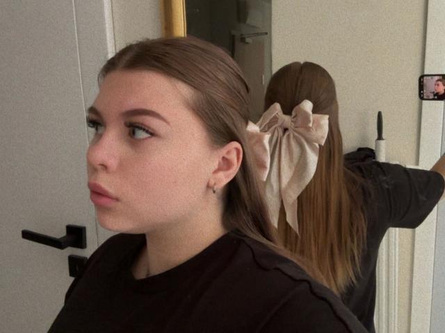 AbbyMaus-hot with brown hair in a black shirt, wearing a large beige bow, looking to the side, reflected in a mirror. Her ass is partially visible.