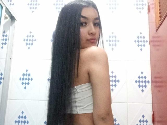 KarinaArisonya with long black hair, looking over her shoulder, wearing a white tube top, showing her bare back and small tits, standing in a tiled bathroom.