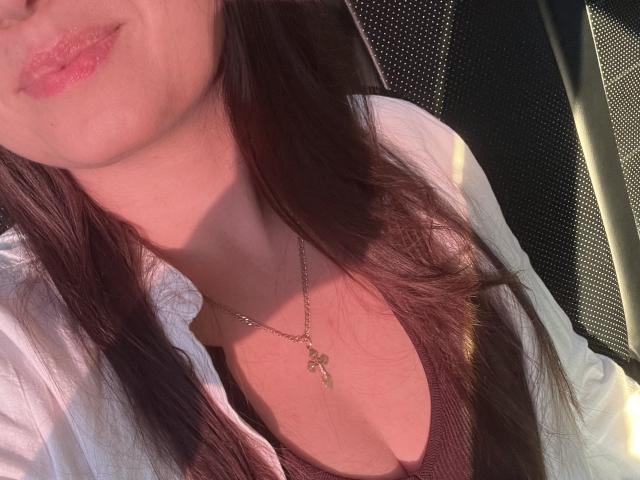 Das Gesicht von AnnaCharm69 mit rosafarbenen Lippen, braunen Haaren, einem weißen Hemd, einem braunen Tanktop und einer goldenen Halskette mit einem Schlüsselanhänger, die ihren Ausschnitt zeigt. Sonnenlicht auf ihrer Haut..