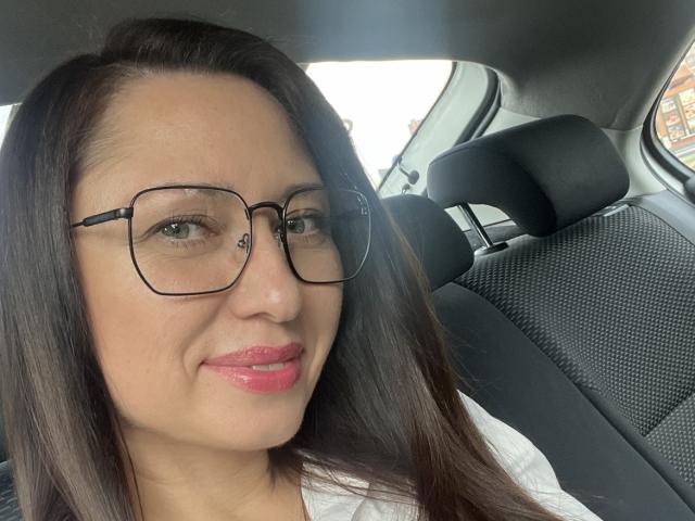 AnnaCharm69, con gafas, lápiz labial rosa y cabello castaño oscuro lacio, sonriendo en un coche. Sus pechos son pequeños y tiene un rostro lindo y atractivo..