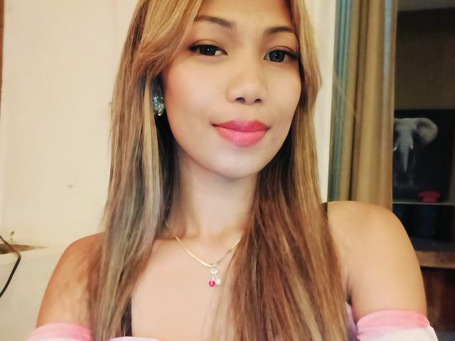 SweetPinkieFrench with long blonde hair, pink lipstick, and a necklace with a pink gem, smiling at the camera, wearing a pink off-shoulder top, showing her tits.