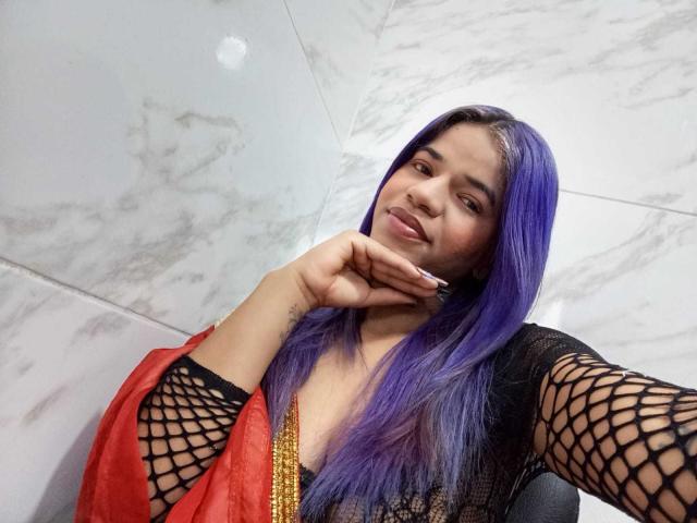 KairaDayamai with purple hair, black fishnet sleeves, and red dress, resting chin on hand, smiling at camera, showing a subtle tattoo on her right hand.