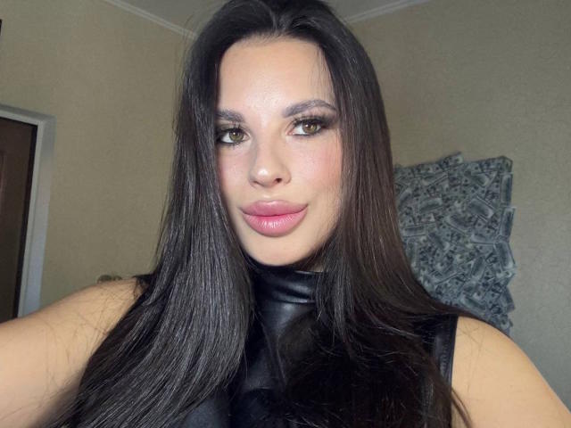 BlackFatal, con lunghi capelli neri e lisci, indossa una giacca di pelle nera, ha gli occhi verdi, le labbra rosa e un leggero sorriso, e guarda direttamente l'obiettivo..