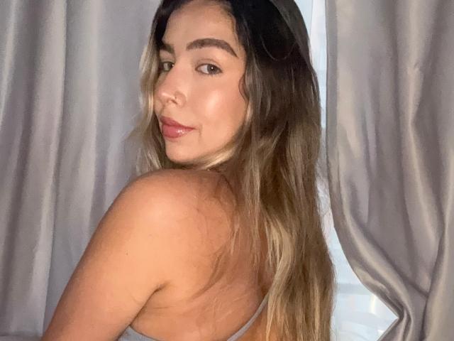 MaraCortez with long brown hair, looking over her shoulder, showing her ass, wearing a gray top, smiling softly, in a dimly lit room with gray curtains.