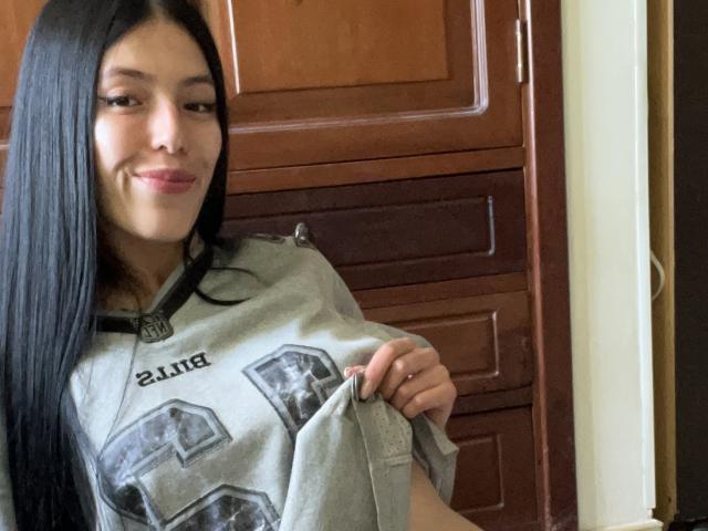 AizenMyoo with long black hair, light skin, and a slight smile, wearing a grey sports jersey, pulling it up to reveal her titty. She's standing in front of wooden doors.