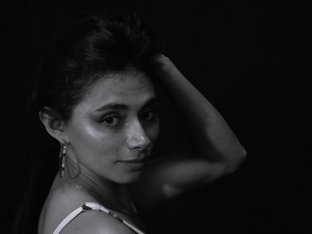 AnnaMooree, avec sa peau éclatante, ses cheveux foncés et ses boucles d'oreilles, regarde de côté, bras levé, portant un haut blanc à bretelles, noir et gris, monochrome, sexy..