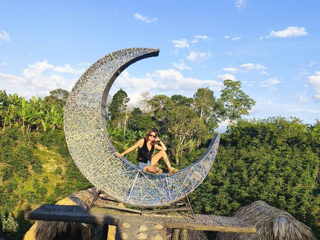 AmelieLautrec is sitting on a large, crescent-shaped mosaic sculpture with blue and yellow tiles, her ass resting on the curved top, surrounded by greenery and a clear sky.
