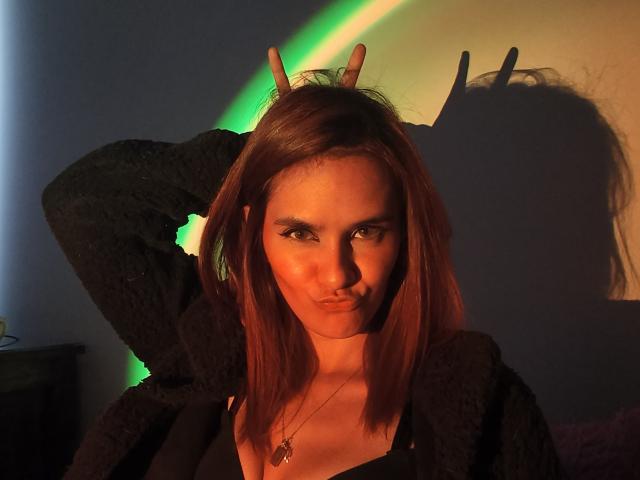 AmelieLautrec with red hair, black sweater, and necklace, making a peace sign with her right hand, shadowed by a green light, has a confident, slightly smug expression.