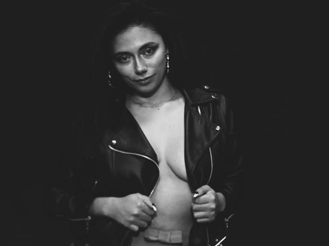 AnnaMooree, foto en blanco y negro, vistiendo una chaqueta de cuero, abierta para revelar sus senos, sosteniendo la chaqueta, fondo oscuro, sonrisa sutil, aretes, cabello oscuro, mirando a la cámara..