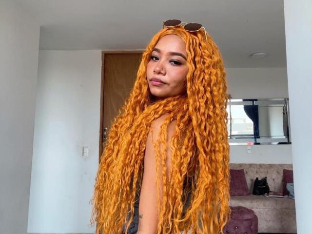 Evakurlly with long, curly orange hair, sunglasses on head, looking over shoulder, slight smile, wearing black sleeveless top, standing in room with white walls and wooden door.