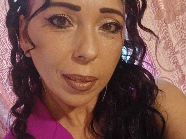 LauraSin has curly black hair, brown eyes, and full pink lips. She's wearing a pink top and has hoop earrings. Her face is slightly tanned with a subtle smile.
