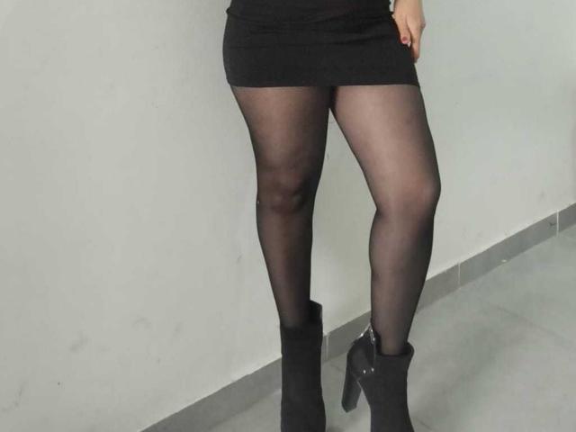 CamileParist's ass is covered by a tight black skirt. She's wearing sheer black pantyhose and black ankle boots with high heels, standing against a white wall.