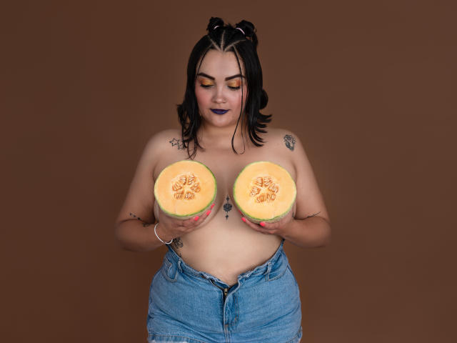 AnthonellaMiller, topless, holding two halves of a pumpkin, has large tits, dark hair in buns, purple lipstick, tattoos, and wears unbuttoned jeans against a brown background.