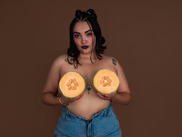 AnthonellaMiller with black hair, purple lipstick, tattoos, holding two halved pumpkins covering her tits, wearing unbuttoned blue jeans, brown background, standing, looking at camera, serious expression.