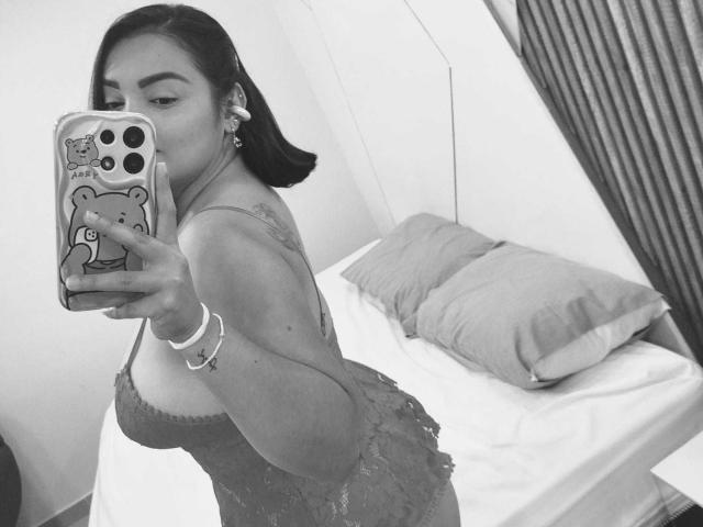 AnthonellaMiller with dark hair in a ponytail, taking a mirror selfie in a lacy dress, showing her ass, in a black-and-white photo. She's holding a phone with a bear case.