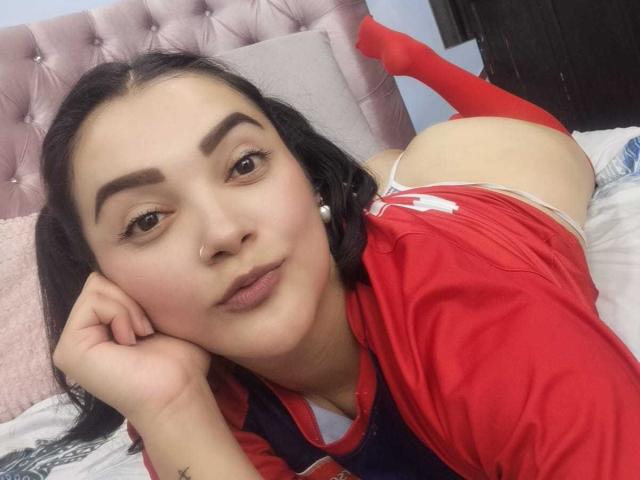 AnthonellaMiller has dark hair, red eyebrows, and is lying on a bed in a red shirt, showing her bare ass in white panties, with red socks on.