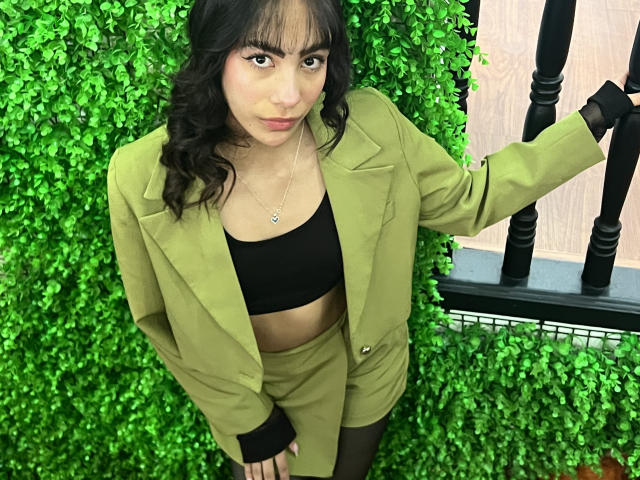 AitanaJohns with dark curly hair, wearing a green blazer, black crop top, and green shorts, standing against a green leafy wall, looking up with a slight pout.