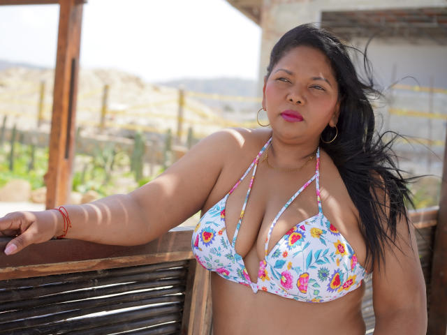 Keilathompson, avec de grandes seins duveteux, porte un bikini floral coloré et se tient debout, un bras appuyé sur une balustrade en bois. Elle a de longs cheveux noirs, du rouge à lèvres rose et des boucles d'oreilles dorées..