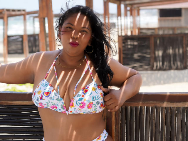 Keilathompson, con i capelli scuri, indossa un bikini floreale, ha un seno prosperoso, rossetto rosso e orecchini a cerchio dorati, è appoggiata a una ringhiera di legno, e la luce del sole proietta ombre..