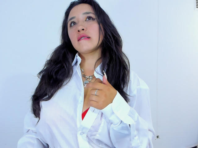 JessyBarnnett with long black hair, wearing a white shirt, holding the collar, showing a red bra, necklace, and ring, smiling with pink lipstick, looking up.
