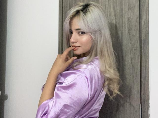 AlexaThoms with blonde hair, purple satin shirt, finger near mouth, looking back, soft smile, sexy, pale skin, slight makeup, wood door behind her, casual, confident, cute.