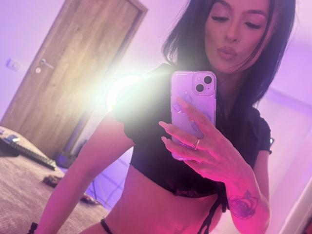 KinkyAmelie with dark hair, pouty lips, wearing black top, showing midriff, holding phone, pinkish-purple lighting, tattoo on right hand, wood door in background, reflection selfie.