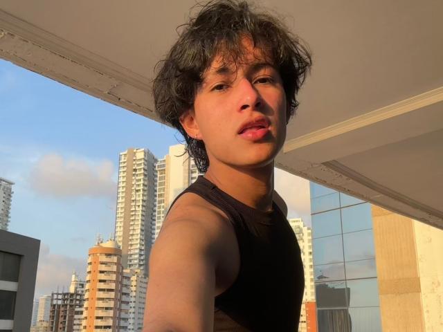 HaroldX mit zerzaustem, schwarzem Haar, der ein schwarzes Tanktop trägt, hat ein süßes Gesicht, leicht geschwollene, rosafarbene Lippen und macht ein Selfie auf einem sonnigen Balkon..