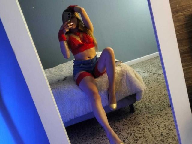 AmbarVelle in red lace bra, denim shorts, and red garter, sitting on white bed, legs crossed, mirror selfie, holding phone, long brown hair, visible ass, and heels.