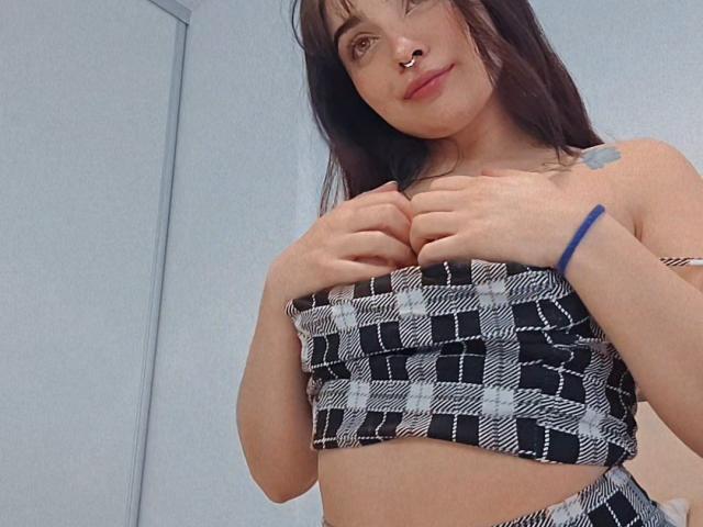 AshleyDaphna with small tits, wearing a black and white checkered tube top, hands covering her chest, nose ring, blue bracelet, and tattoo on her right shoulder, smiling.