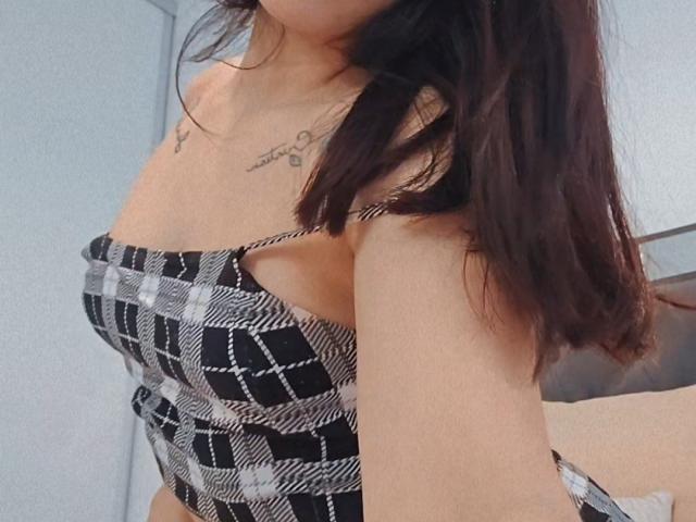 AshleyDaphna, con i capelli castano scuro, indossa una maglietta a quadri bianchi e neri, che mette in mostra piccoli seni e un tatuaggio sul petto, con le braccia leggermente alzate, in un ambiente interno informale..