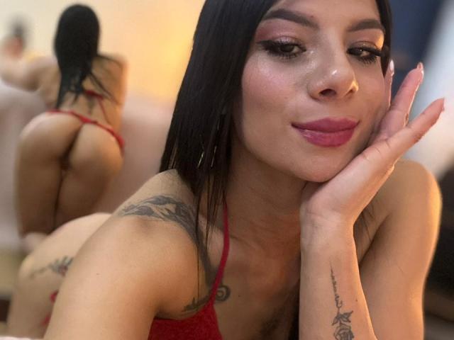MelissaSpencer with long black hair, red lipstick, and tattoos, wearing red lingerie, smiling at the camera, hand on cheek, ass and back in mirror reflection, bedroom.