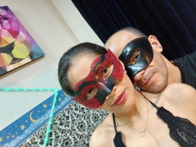 CoupleHot69s with glittery red mask, small tits, black lace top, and CoupleHot69s with black mask, bald head, black shirt, nuzzling her neck, both smiling.
