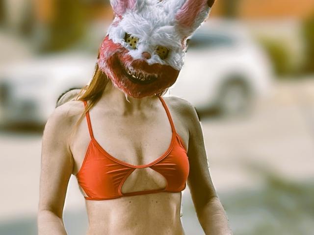 Una ragazza attraente con una maschera da coniglio bianca e rosa, indossa un bikini rosso con un taglio, che mostra piccoli seni e un ventre piatto, in piedi all'aperto..